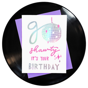 the words go shawty it's your birthday in shiny holographic rainbow foil with pink accents & a shiny disco ball for the o on top of a purple envelope. block printing by Niki Baker with foreignspell from hand-carved stamps & song lyrics.